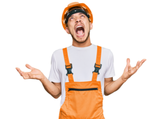 Hispanic young man wearing handyman uniform and safety hardhat crazy and mad shouting and yelling with aggressive expression and arms raised. frustration concept.