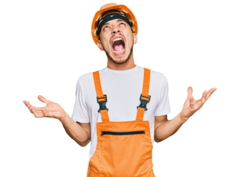 Hispanic young man wearing handyman uniform and safety hardhat crazy and mad shouting and yelling with aggressive expression and arms raised. frustration concept.
