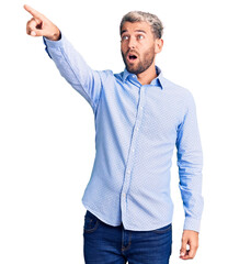Young handsome blond man wearing elegant shirt pointing with finger surprised ahead, open mouth...