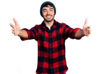 Hispanic young man with beard wearing wool winter hat looking at the camera smiling with open arms for hug. cheerful expression embracing happiness.