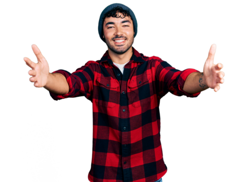 Hispanic young man with beard wearing wool winter hat looking at the camera smiling with open arms for hug. cheerful expression embracing happiness.