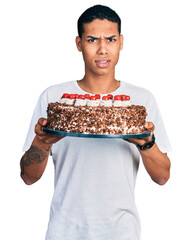 Young hispanic man celebrating birthday holding big chocolate cake clueless and confused...