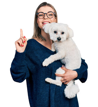 Young blonde woman hugging cute dog smiling with an idea or question pointing finger with happy face, number one