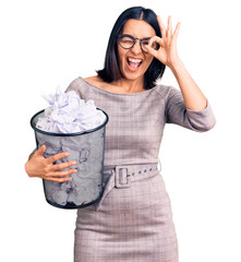 Young beautiful latin woman holding paper bin full of crumpled papers smiling happy doing ok sign with hand on eye looking through fingers