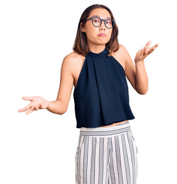 Young beautiful chinese girl wearing casual clothes and glasses clueless and confused expression with arms and hands raised. doubt concept.