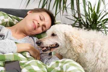 Throughout the day, a middle-aged woman with short hair relaxes in bed with her large poodle....