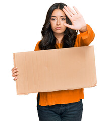 Beautiful brunette young woman holding cardboard blank empty banner with open hand doing stop sign...