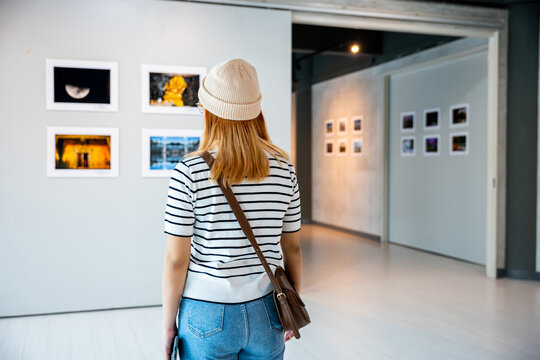 Asian young woman standing she looking art gallery in front of colorful framed paintings pictures on white wall, female watch at photo frame to leaning against at exhibit museum, Back view