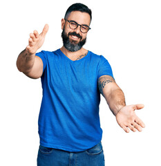 Hispanic man with beard wearing casual t shirt and glasses looking at the camera smiling with open arms for hug. cheerful expression embracing happiness.