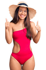 Young beautiful latin girl wearing swimwear and summer hat success sign doing positive gesture with hand, thumbs up smiling and happy. cheerful expression and winner gesture.