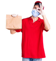 Young hispanic man wearing delivery uniform and medical mask holding paper bag smiling happy doing ok sign with hand on eye looking through fingers