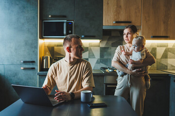 A young couple with a child in the kitchen. A man works at home on a laptop, and his wife with a...