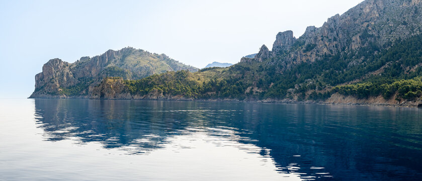Morning serenity of Mallorca coastline, bathed in sunlight and veiled in gentle haze, featuring Morro des Forat headland and Cingle d'en Perot cliff with Torre de Sa Mola de Can Palou watchtower.