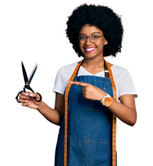 Young african american woman dressmaker designer wearing atelier apron holding scissors smiling...