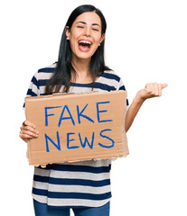Beautiful young woman holding fake news banner screaming proud, celebrating victory and success...