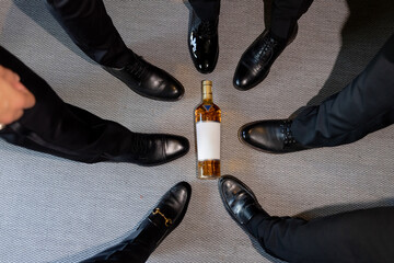 Men's party with alcohol indoors. Men in classic shoes close-up in a circle and a bottle of whiskey...