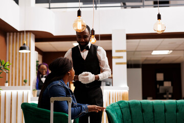 Smiling friendly hotel bellboy wearing uniform talking with African American woman guest sitting in...