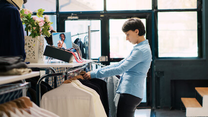 Store employee arranging hangers full with stylish clothes, working at modern boutique visual....