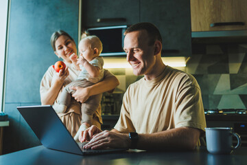 A young father works on a laptop while his wife takes care of the child. Work from home. A young...