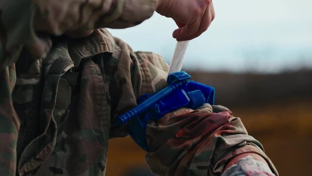 Close-up of a confident male soldier in a green camouflage uniform puts a blue tourniquet on his arm to avoid blood loss when wounded during first aid training at a military training ground