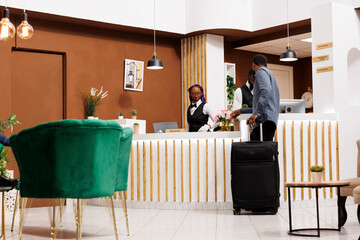 Hotel check in process. African American man traveler with suitcase standing at reception desk...