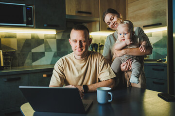 A young father works on a laptop while his wife takes care of the child. Work from home. A young couple in the kitchen with a child