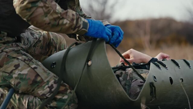 From the side close up a confident male army medic in camouflage green clothing and blue rubber gloves ties straps on a stretcher to move a wounded soldier during combat operations in the steppe