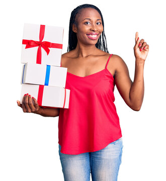 Young african american woman holding gift surprised with an idea or question pointing finger with happy face, number one