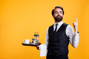 Waiter with serving tray points upwards on camera, showing something up above head while he wears professional restaurant clothes. Young man butler suggesting direction in studio. - Powered by Adobe