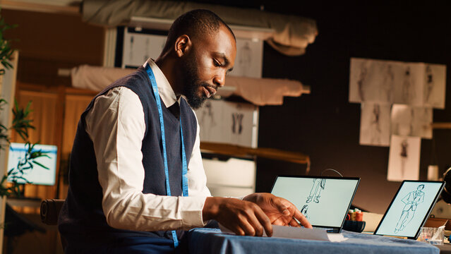 Tailor reviewing designs on tablet, comparing sketches of clothes in atelier workshop. Fashion couturier manufacturing clothing line using tailoring instruments and tools. Handheld shot.