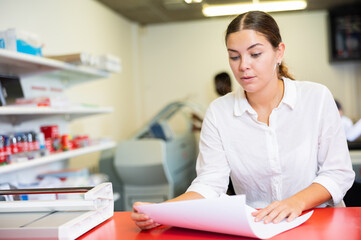 Focused young woman in white shirt checking the print quality and accepting color proofs in...