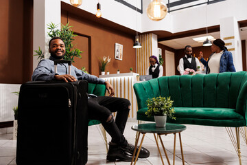 Happy smiling African American man guest sitting in hotel lobby with suitcase awaiting for room....