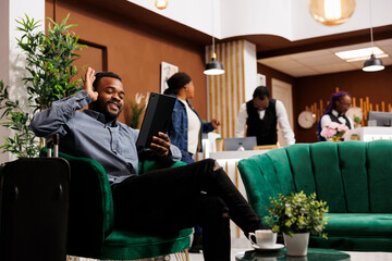 Happy smiling young African American man traveler sitting in hotel lobby having video call while...
