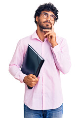 Handsome young man with curly hair and bear holding business folder serious face thinking about...