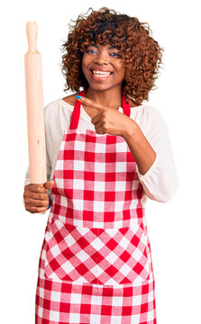 Young african american woman wearing apron holding rolling pin smiling happy pointing with hand and finger
