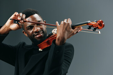 Elegant African American musician playing violin in black suit on gray background in studio setting