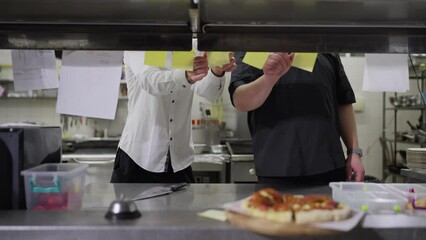 A male waiter in a white shirt and a cook in a black uniform work with stickers with orders from the hall in the kitchen in a restaurant. A male cook together with a waiter places and inspects all