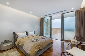 A neatly arranged bedroom boasts a large comfortable bed with beige bedding, facing glass sliding doors that reveal a tranquil ocean view.