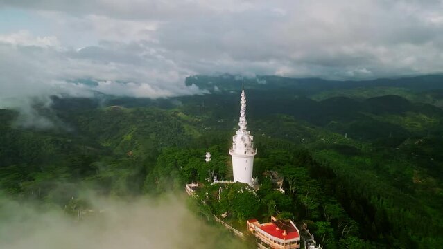 Drone sweeps over Ambuluwawa Tower, Sri Lanka, mist hugging green mountains. Sacred architecture stands tall, peace, unity diverse religions. Visitors climb, explore multi-faith monument, views.