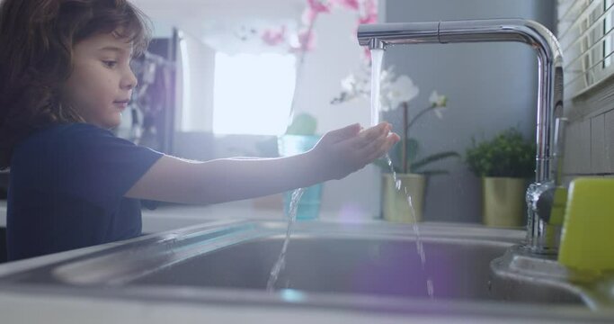 A curious young boy is closely observing the stream of water flowing from a modern kitchen faucet, with a focus on the clear, running water.