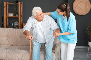 Obraz premium Nurse helping senior man with stick to stand up from sofa at home