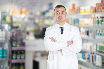 Positive young male pharmacist posing in chemist's shop with large assortment of medicinal...
