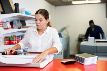 Positive female employee at printing house cuts paper with a paper cutter