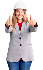 Beautiful young woman wearing architect hardhat approving doing positive gesture with hand, thumbs up smiling and happy for success. winner gesture.