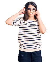 Brunette teenager girl wearing casual clothes and glasses covering ears with fingers with annoyed expression for the noise of loud music. deaf concept.