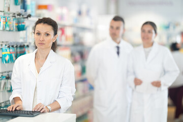Positive middle-aged female pharmacist standing at the desk using keyboard in chemist's shop