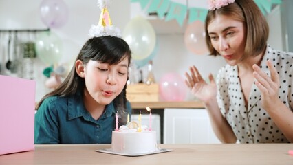 Happy cute child making a wish and blowing birthday cake candle surrounded by family wearing party...