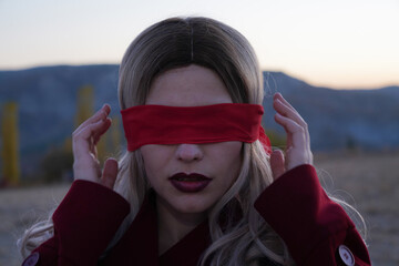 A woman wearing a red coat and a red blindfold. She is standing in a field. Scene is mysterious and...