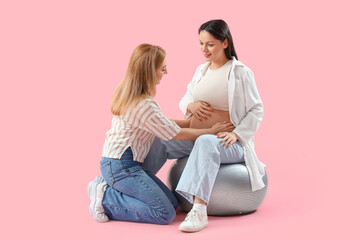 Mature doula and pregnant woman sitting on fitball against pink background