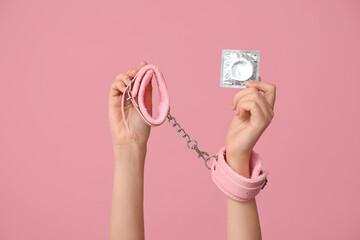 Female hands with handcuffs from sex shop and condom on pink background, closeup